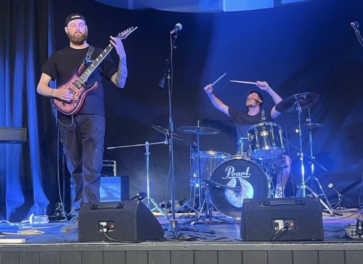 Musicians performing on stage; one playing guitar and the other on drums.
